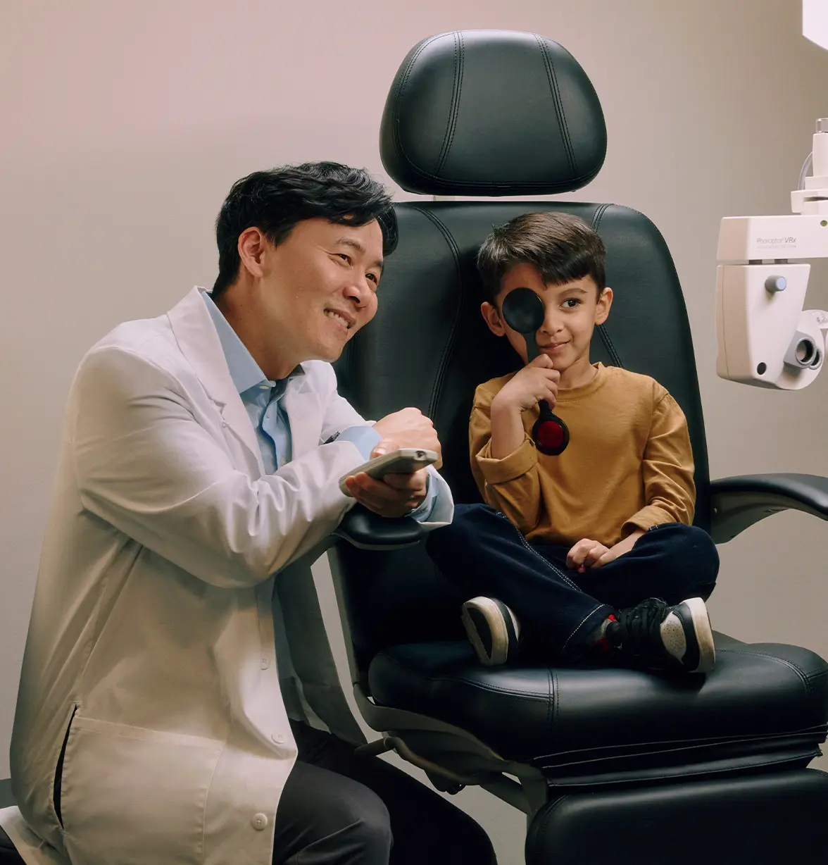 An optometrist smiles beside a young boy, seated in an exam chair, holding a occluder during an eye test.
