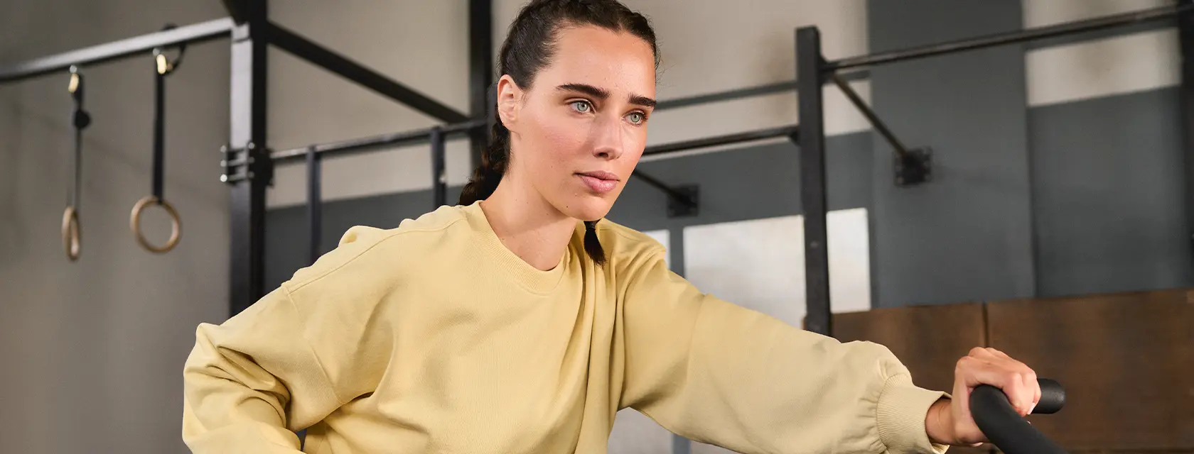 A woman with braided hair, wearing a yellow sweatshirt and green pants, intensely exercises on a stationary bike in a gym.
