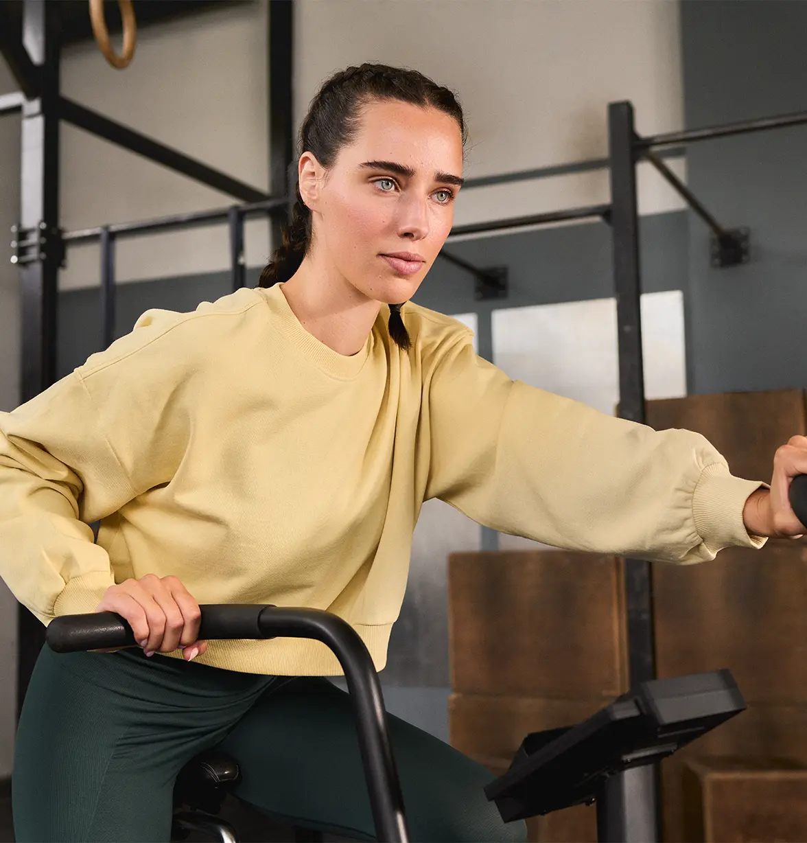 A woman with braided hair, wearing a yellow sweatshirt and green pants, intensely exercises on a stationary bike in a gym.