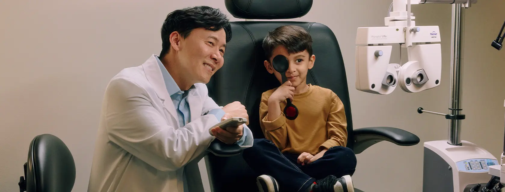 An optometrist smiles beside a young boy, seated in an exam chair, holding a occluder during an eye test.