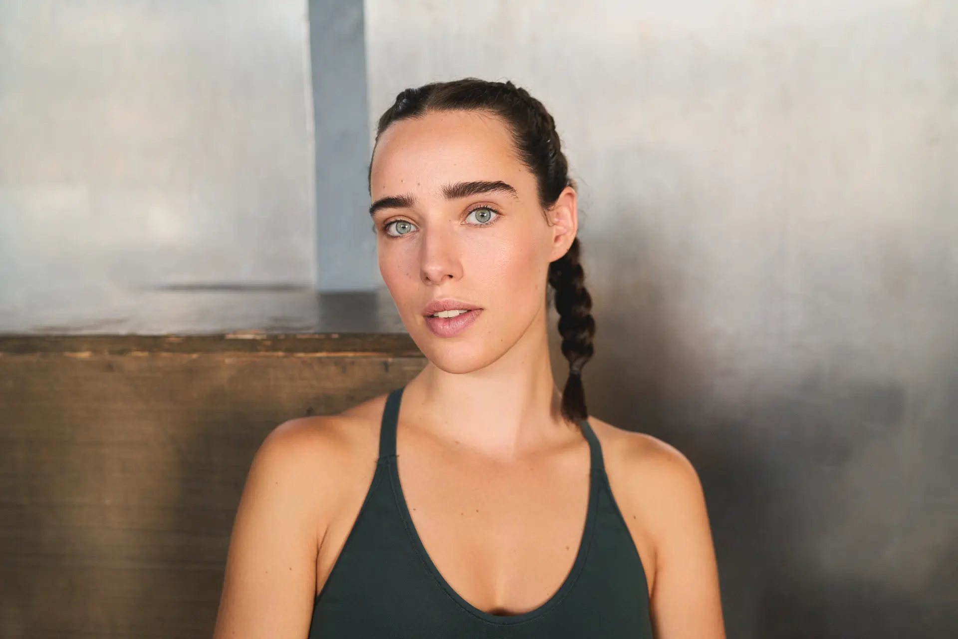 A close-up portrait of a person in athletic wear looking directly at the camera. Their hair is tied back in braids, and the softly lit indoor setting creates a calm, focused atmosphere that suggests a break before or after exercise.