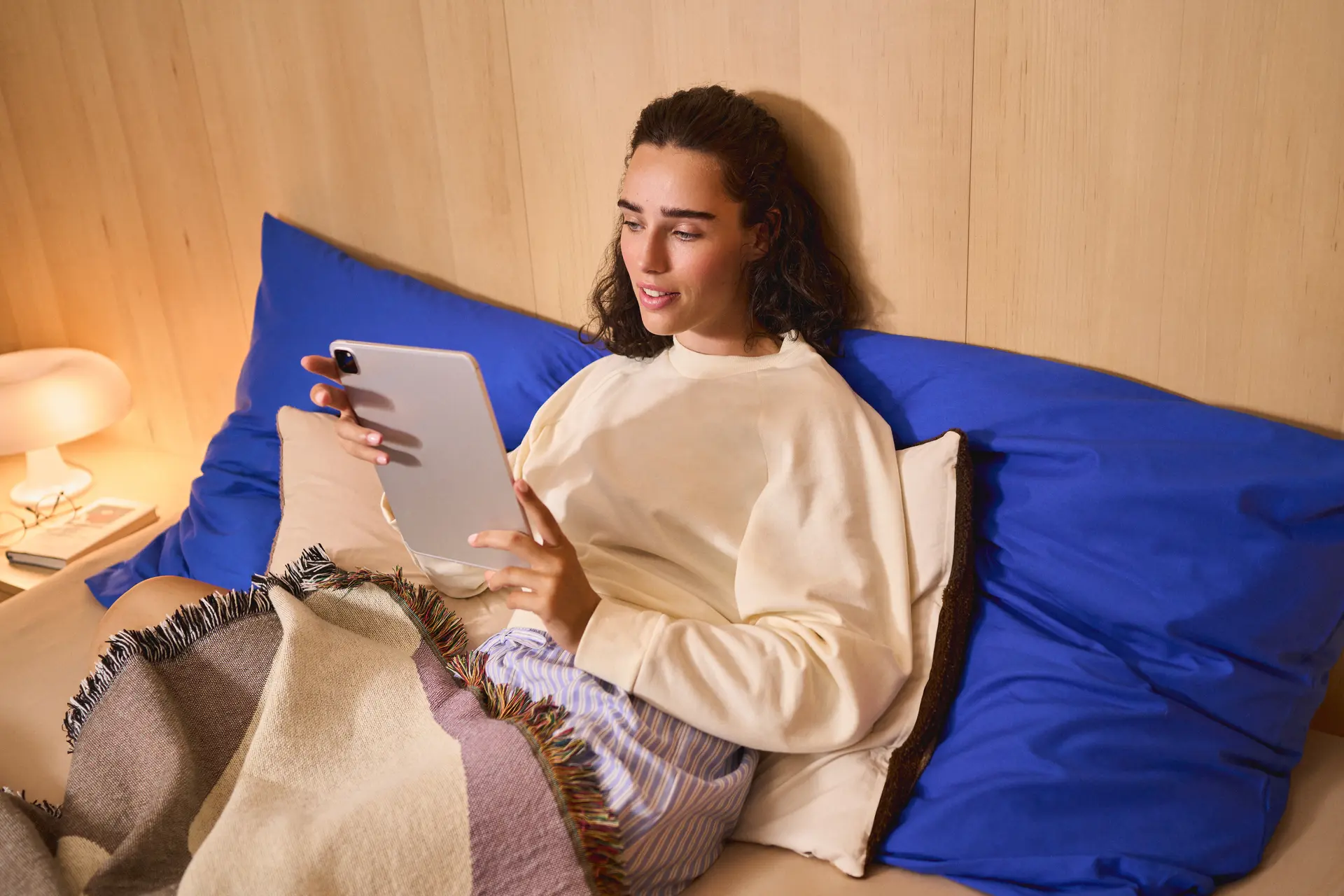 A person is relaxing in bed, leaning against large bright blue pillows while using a tablet. They are wearing a cream-colored sweatshirt and are partially covered by a striped blanket in muted beige, purple, and gray tones with fringed edges. The room has a warm, cozy atmosphere with a light wood-paneled wall behind the bed. On the bedside table, there is a softly glowing mushroom-shaped lamp, a pair of eyeglasses, and a couple of books. The person appears focused on the tablet, suggesting a calm evening of reading, browsing, or streaming content.