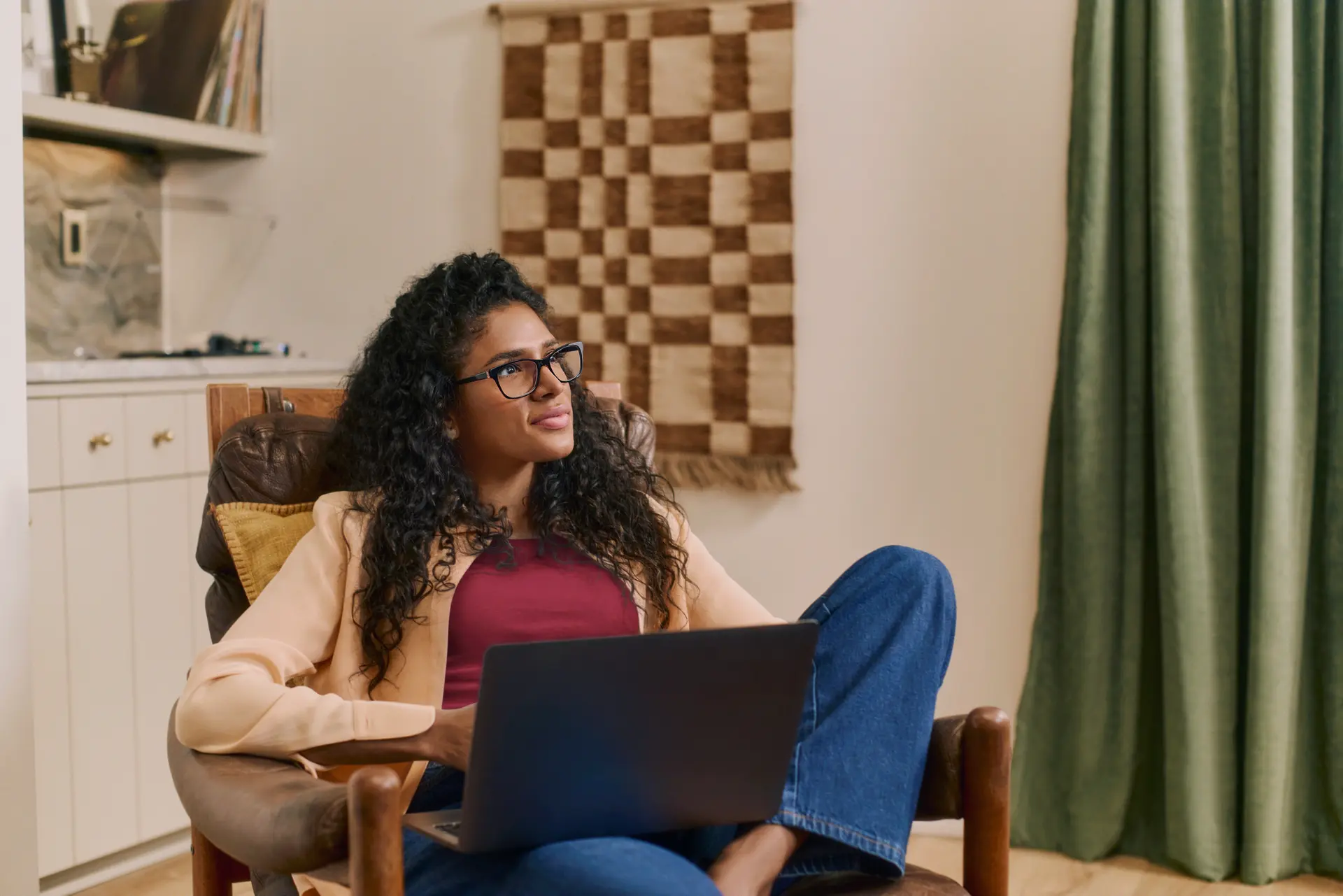 A woman wearing dark-framed glasses sits curled up in a leather armchair with a laptop on her lap. She looks thoughtfully to the side in a cozy room with shelves, a woven wall hanging, and green curtains.