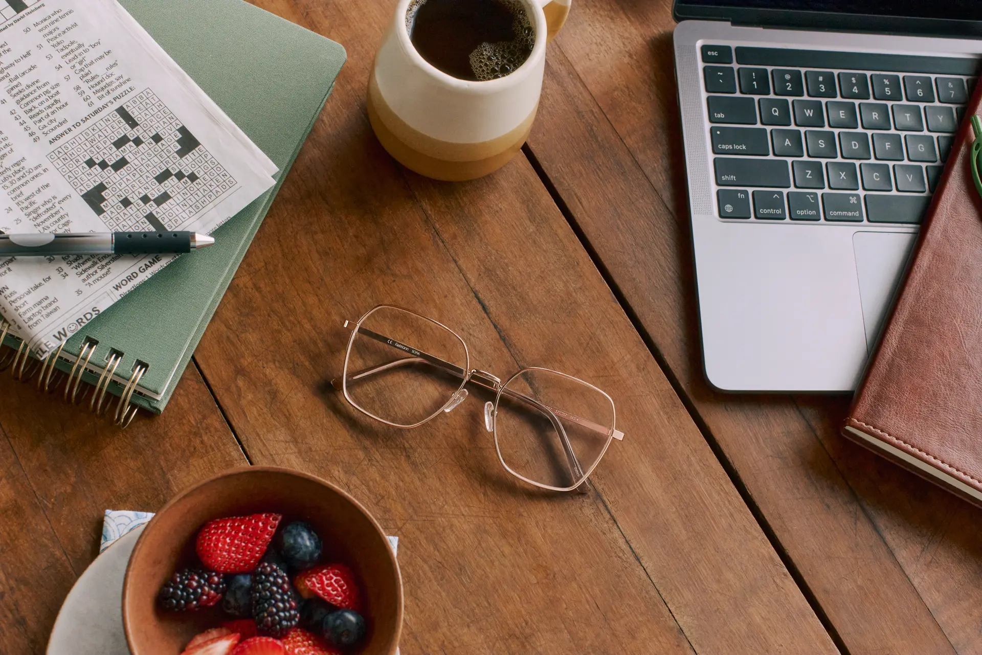 A pair of geometric metal eyeglasses rests on a wooden table surrounded by a laptop, a notebook, a crossword puzzle, a mug of coffee, and a bowl of fresh berries. The neatly arranged workspace creates a calm and productive home-office atmosphere.