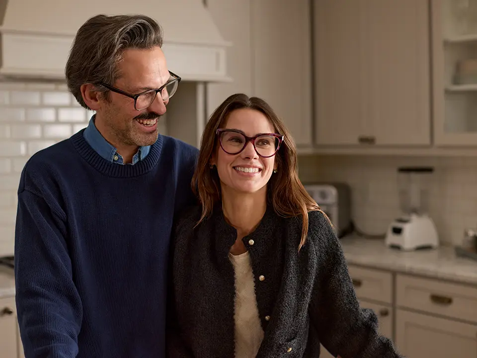 A smiling couple with glasses stands in a kitchen, leaning against the countertop. The man is wearing a blue sweater, and the woman a dark jacket.