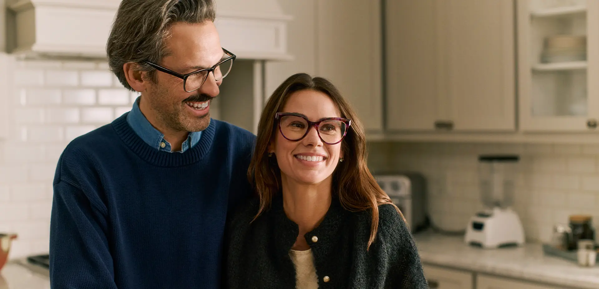 A smiling couple wearing glasses stands closely together in a modern kitchen with white cabinets and a tiled backsplash.