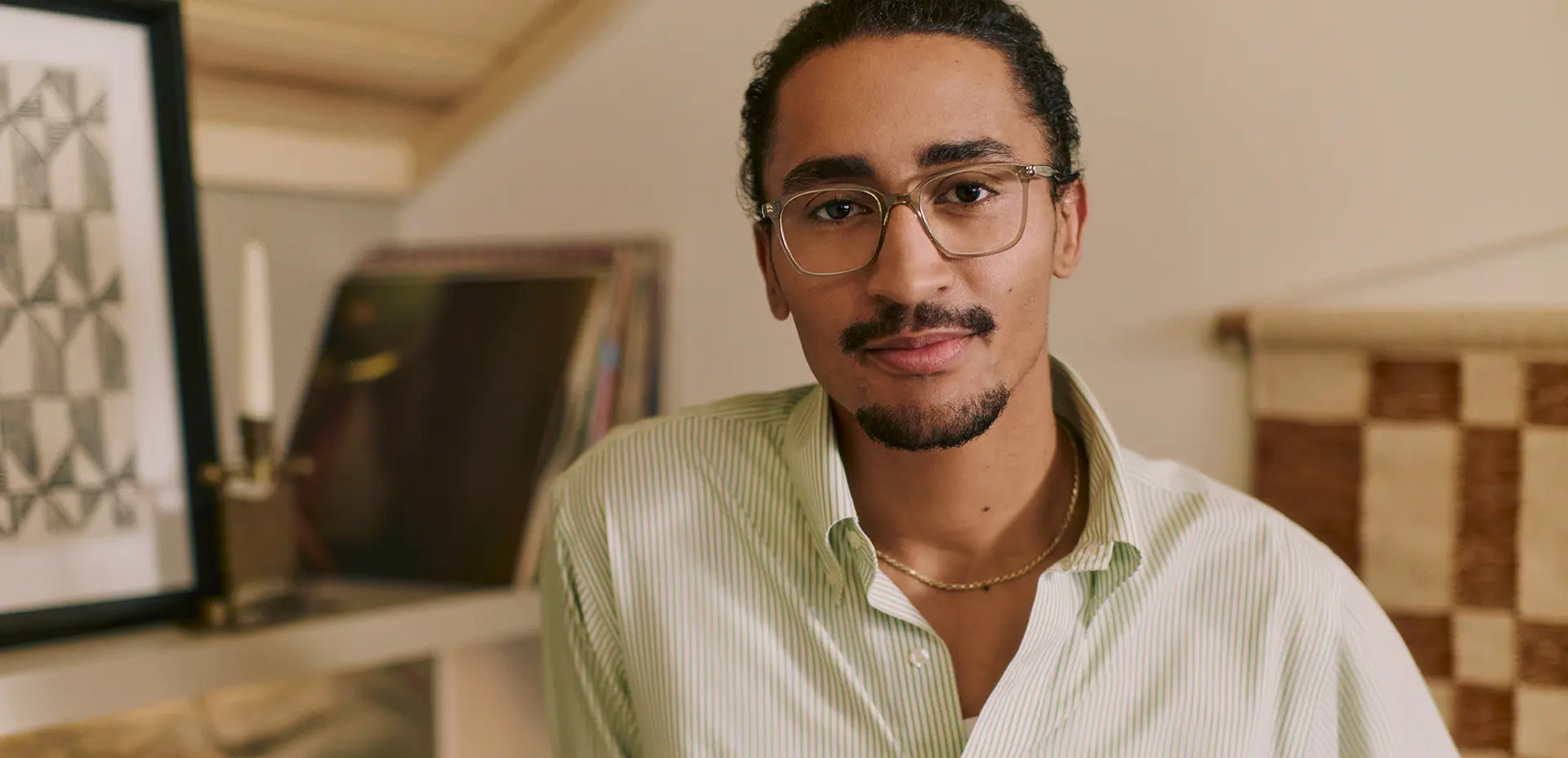 A man wearing glasses and a striped shirt stands indoors, looking at the camera, with art and decorative items in the background.