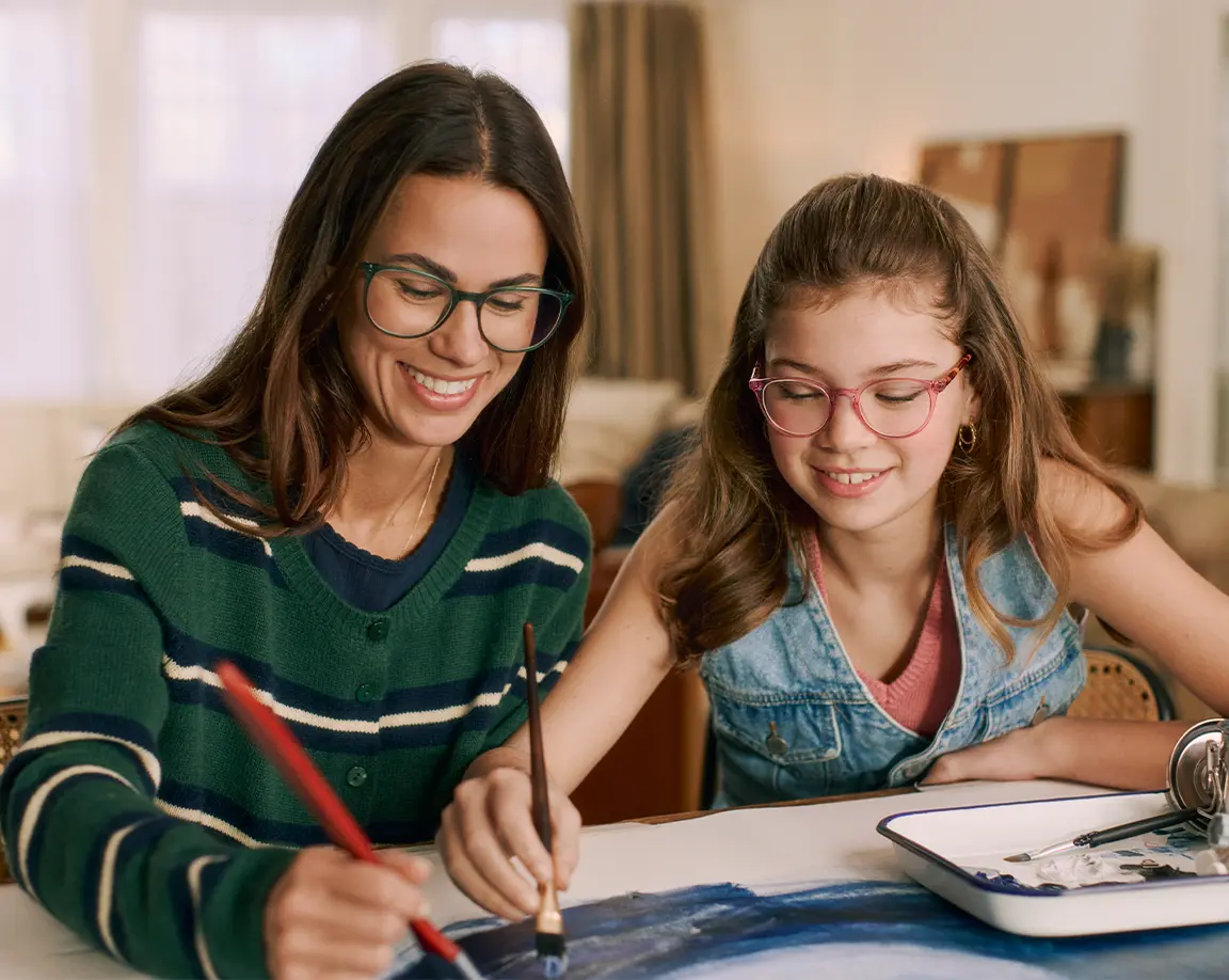 A woman and a girl, both wearing glasses, smile while painting together at a table, with art supplies visible nearby.
