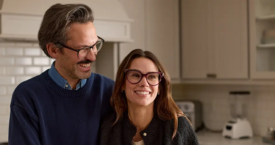 A smiling couple with glasses stands in a kitchen, leaning against the countertop. The man is wearing a blue sweater, and the woman a dark jacket.
